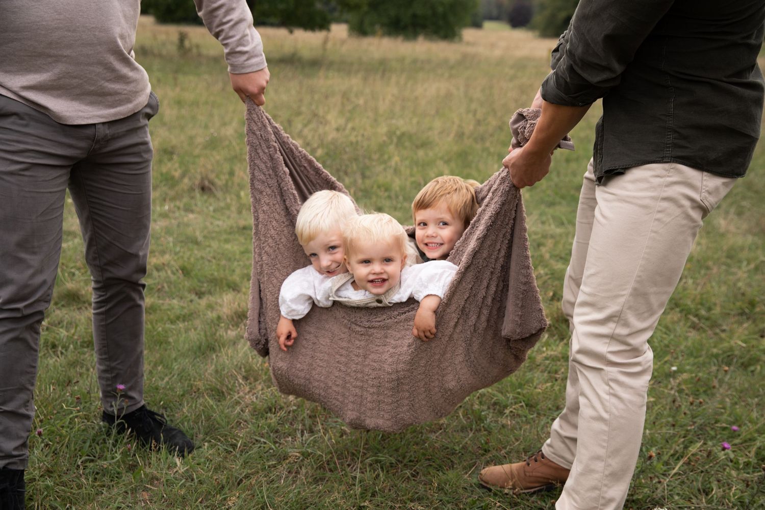 Two children being swung in a brown blanket hammock by adults in a grassy field.