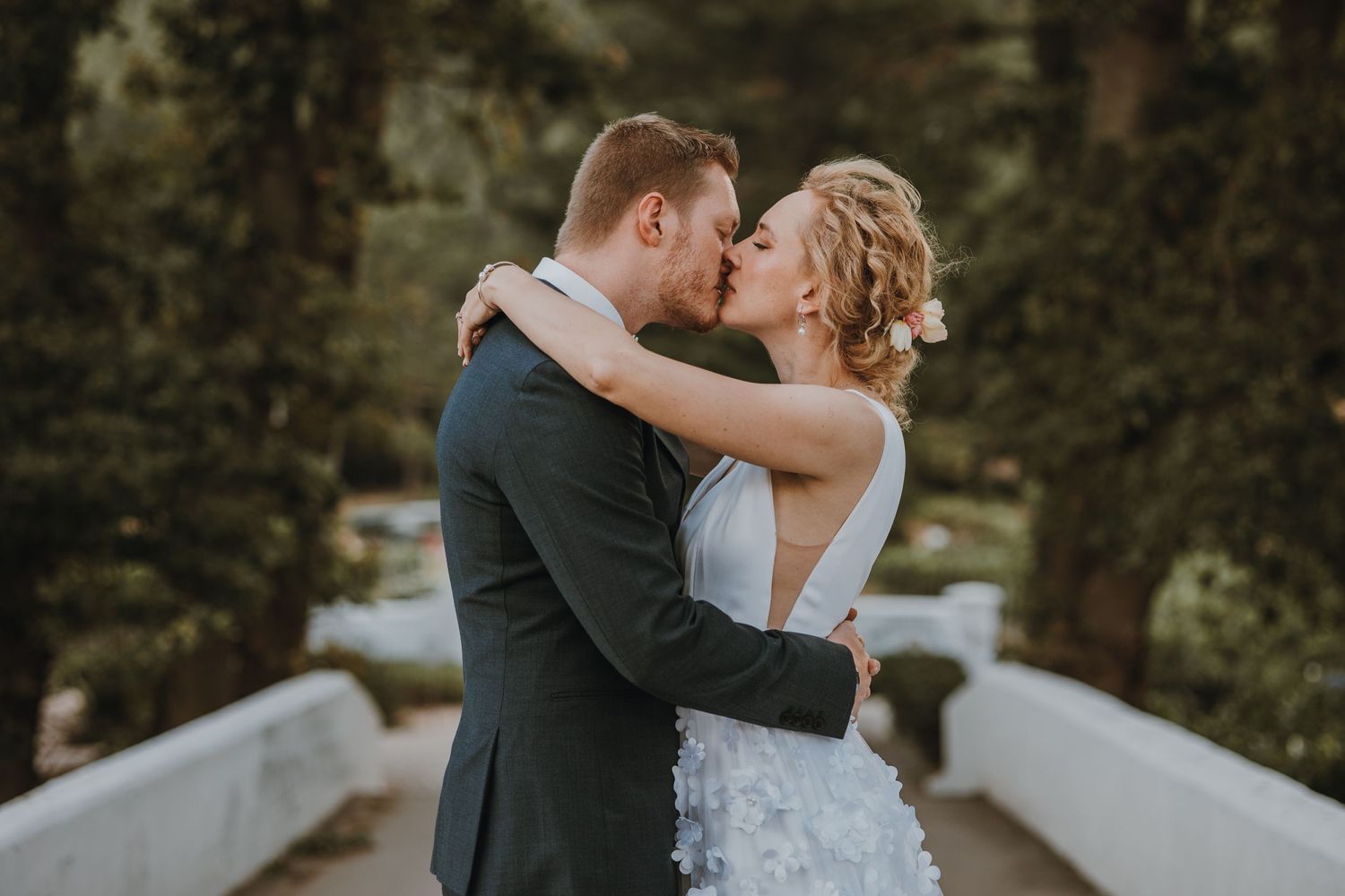 A romantic couple in wedding attire embrace and kiss on a rustic bridge surrounded by greenery.