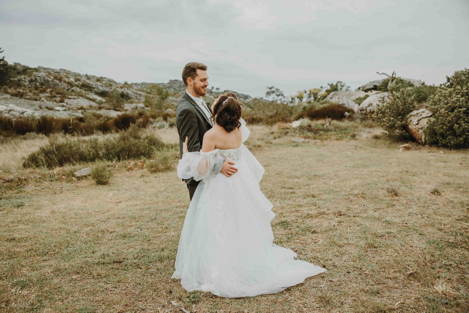 A romantic couple in wedding attire embrace and kiss on a rustic bridge surrounded by greenery.