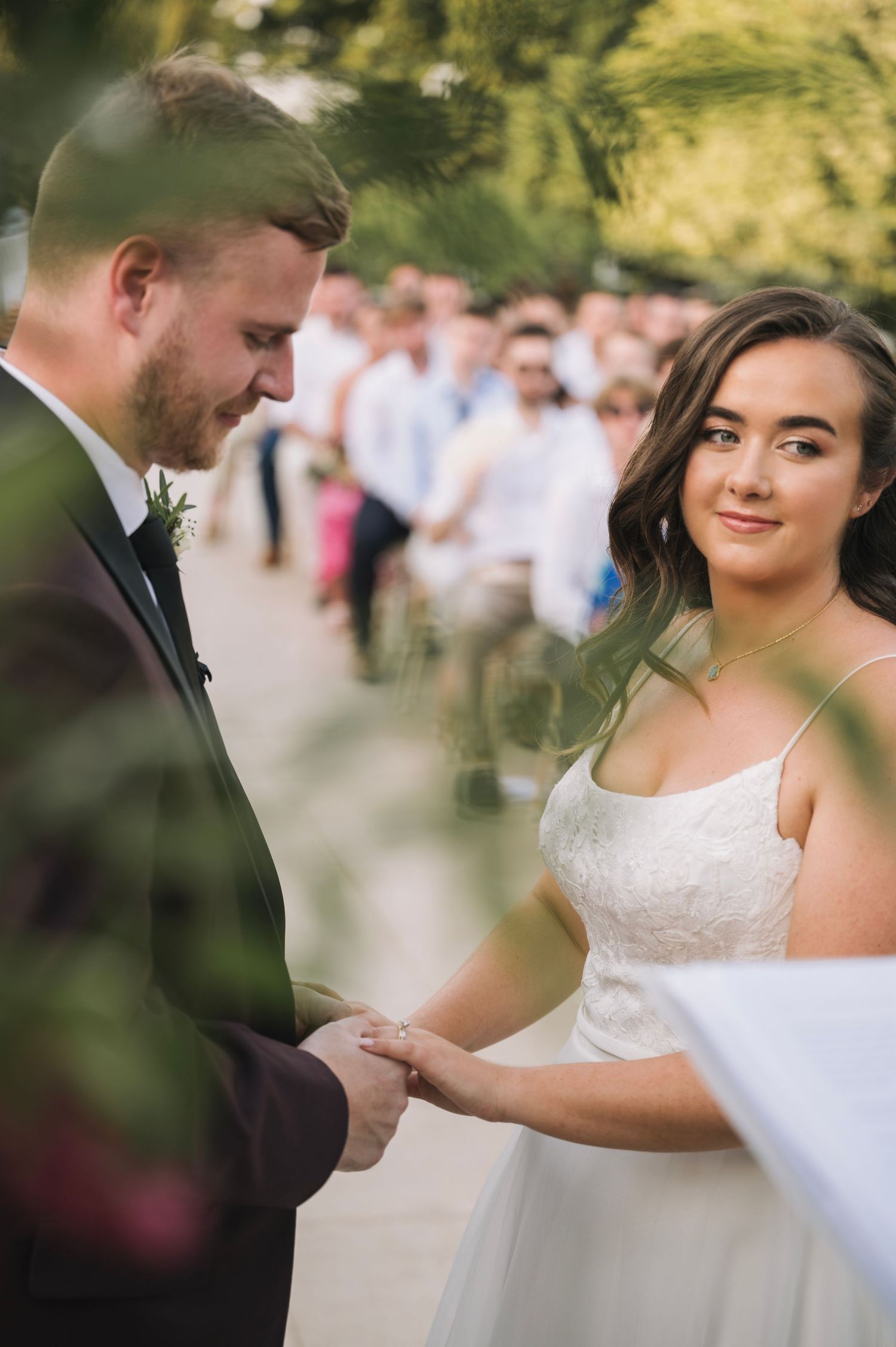 Two people in wedding attire share an intimate moment during outdoor ceremony with guests in background.