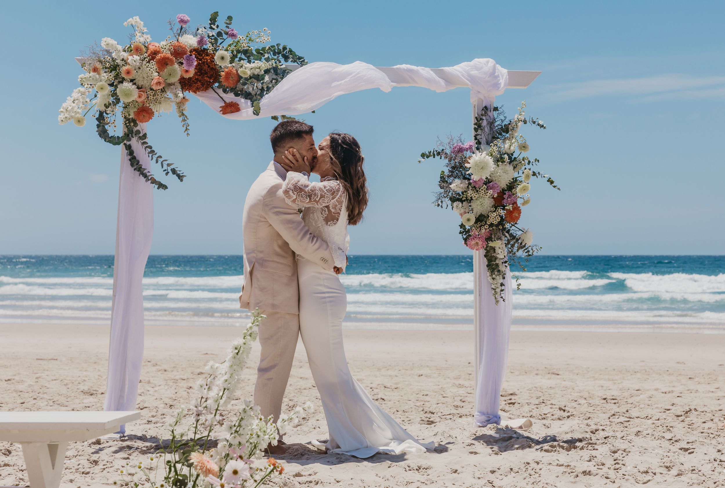 Bride and groom sharing their first kiss beneath a floral wedding arbour on a Byron Bay beach.