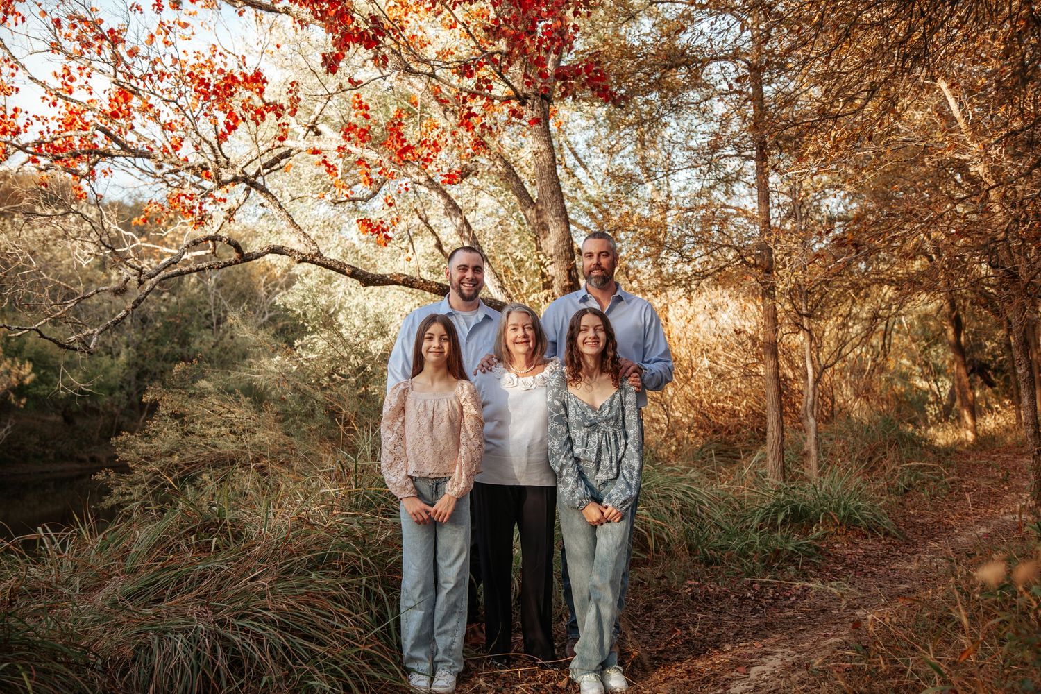 A family: mom, brothers, and granddaughters, stands together under autumn trees with golden foliage along a wooded trail during sunset.