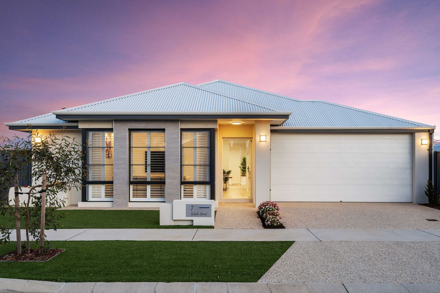 Modern single-story house exterior with white walls and large garage door at sunset with pink and purple sky.