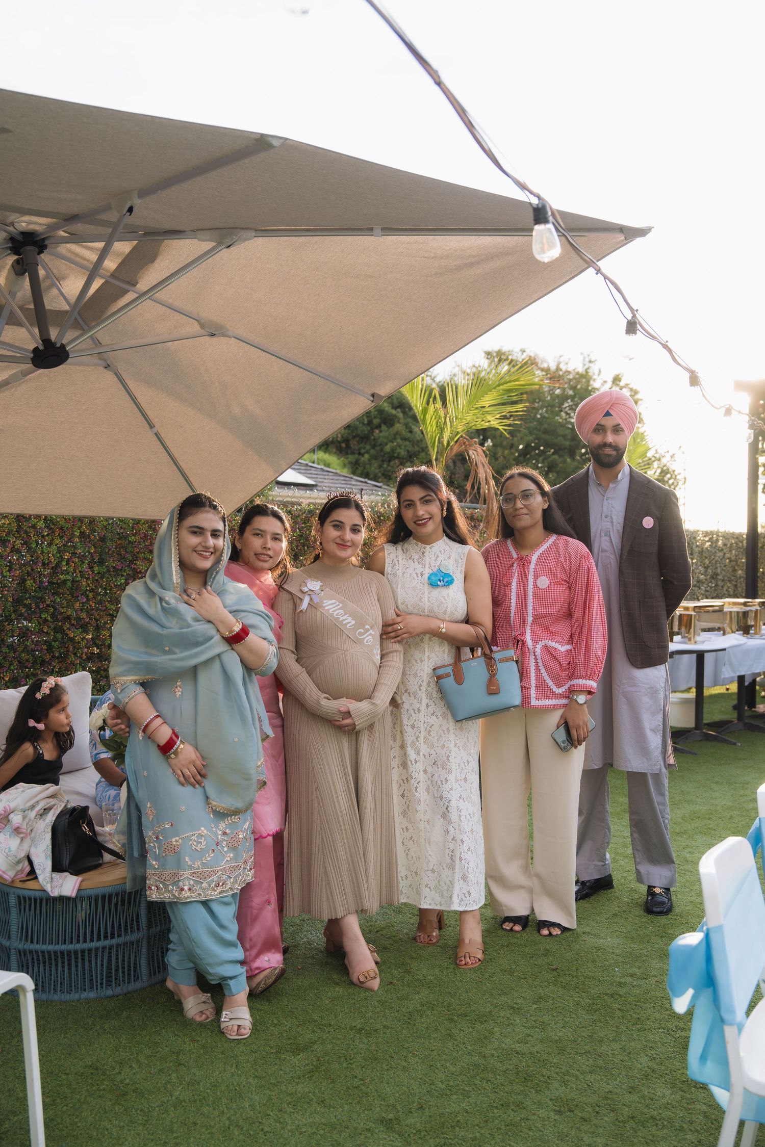 A group gathers under white canopies on a lawn for an outdoor celebration wearing pastel and neutral colored attire.