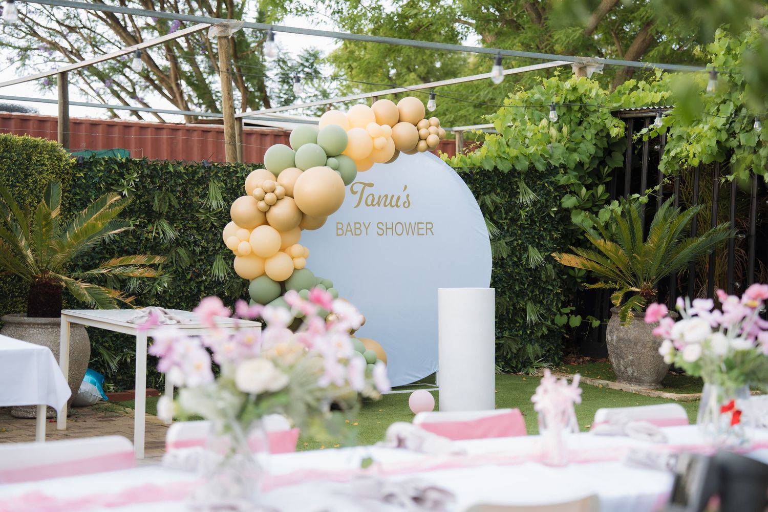 Elegant outdoor party setup with blush pink and gold balloon garlands decorating white tables and chairs under trees.