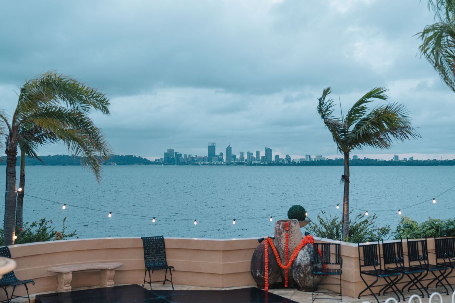 Palm trees sway in the wind along a waterfront promenade with a city skyline visible in the distance across the bay.