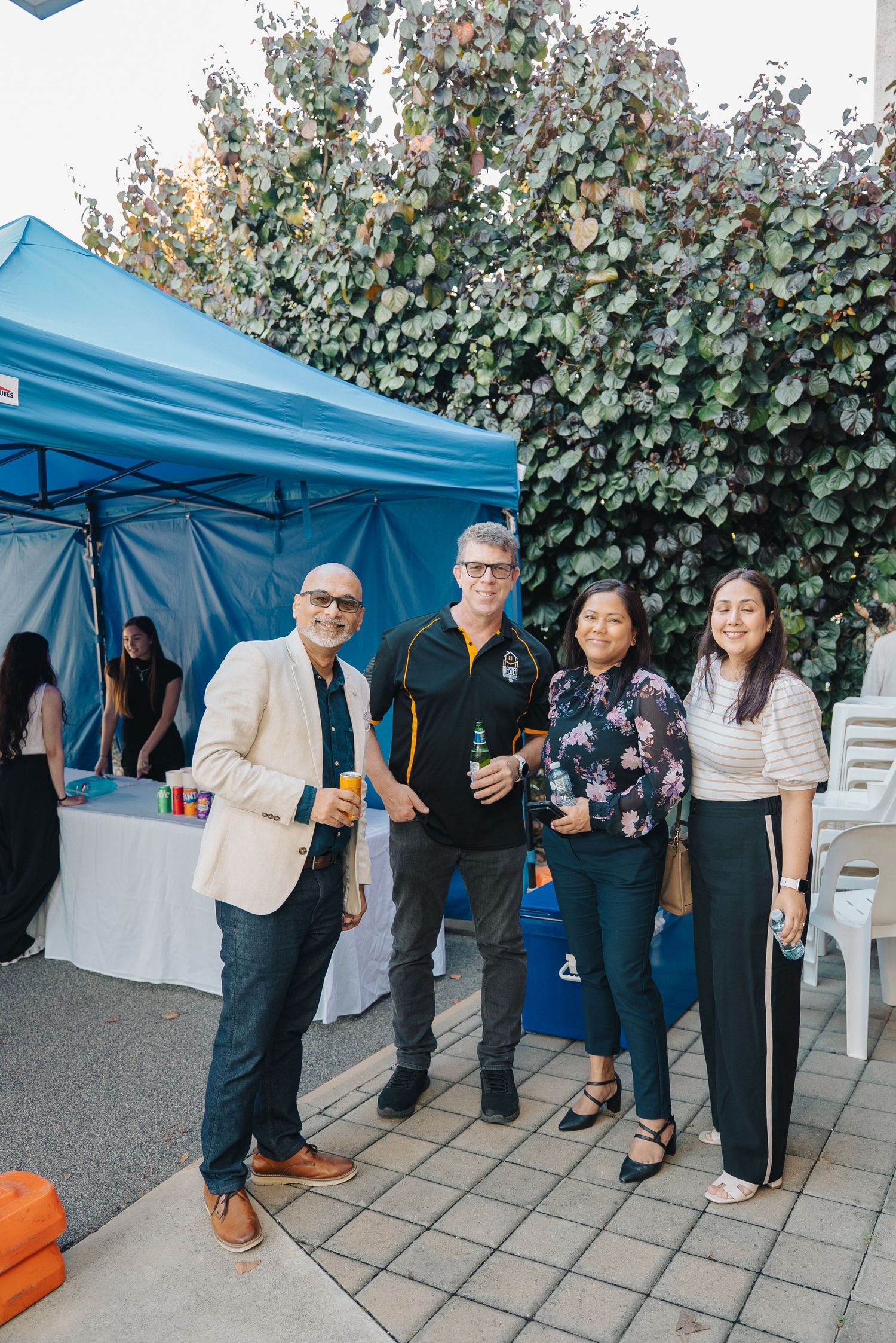 A group of people gathered at an outdoor event under a blue pop-up tent with greenery in the background.