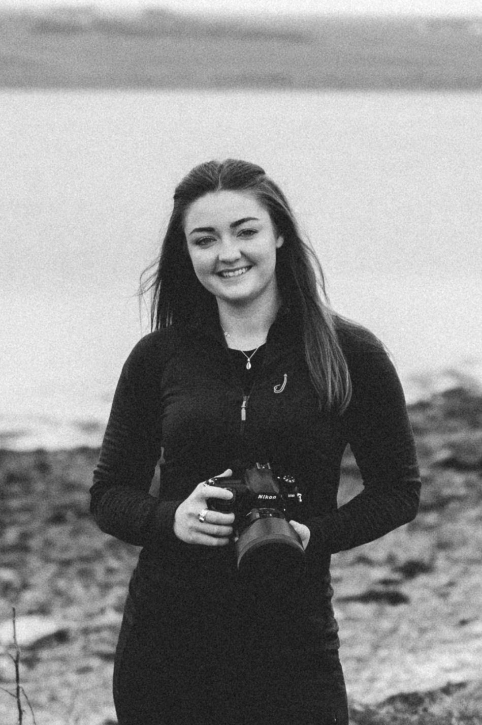 A photographer in a black sweatshirt holds a camera while standing on a rocky beach in black and white.