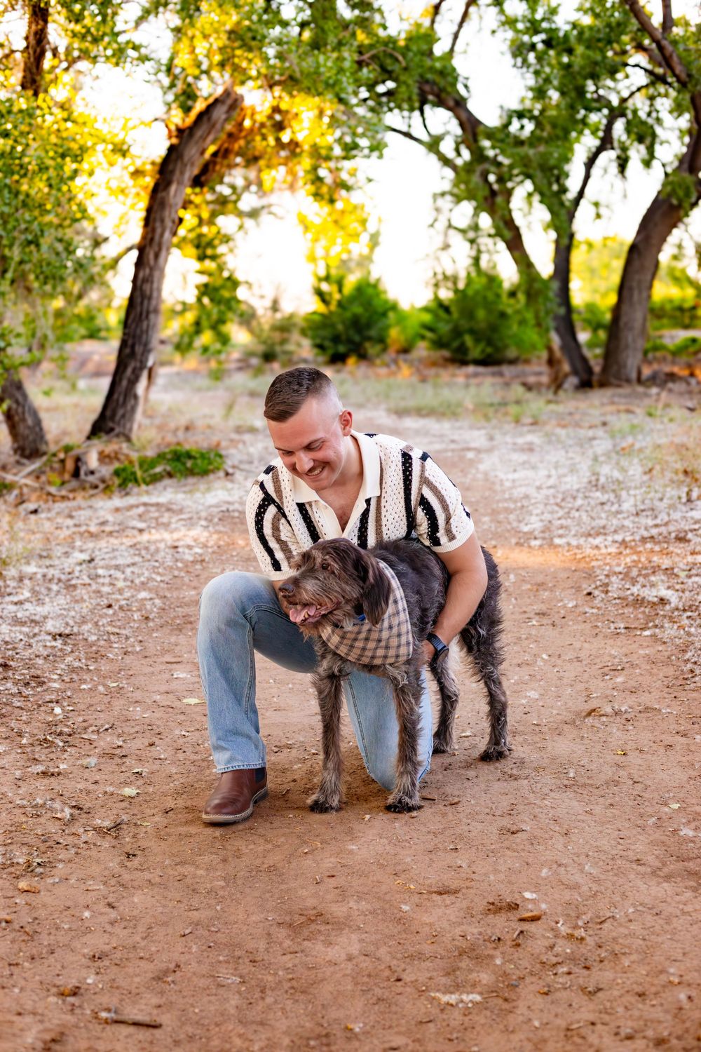 Happy person in striped shirt and jeans petting a large grey dog on a dirt path surrounded by trees during sunset.