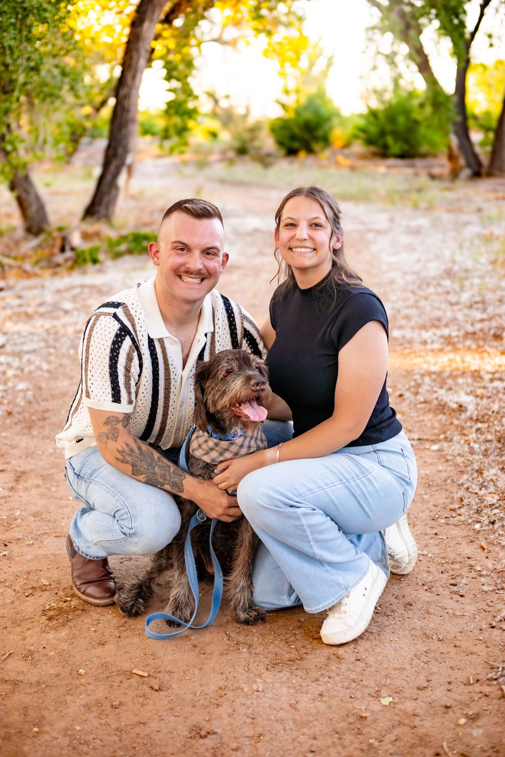 A couple poses outdoors with their small brown dog on a dirt path surrounded by trees on a sunny autumn day.
