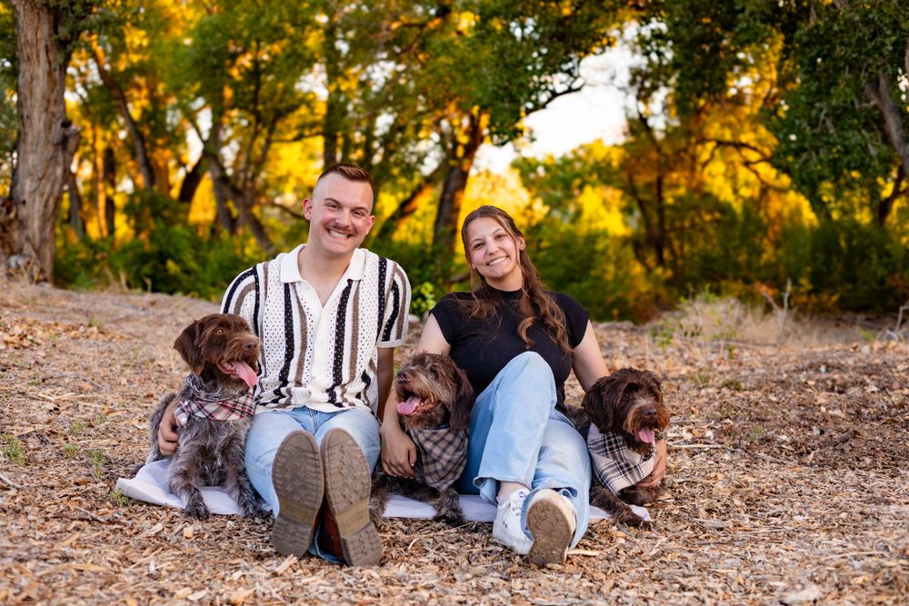Two people sitting outdoors with three Australian Shepherd dogs during golden sunset in a wooded area.