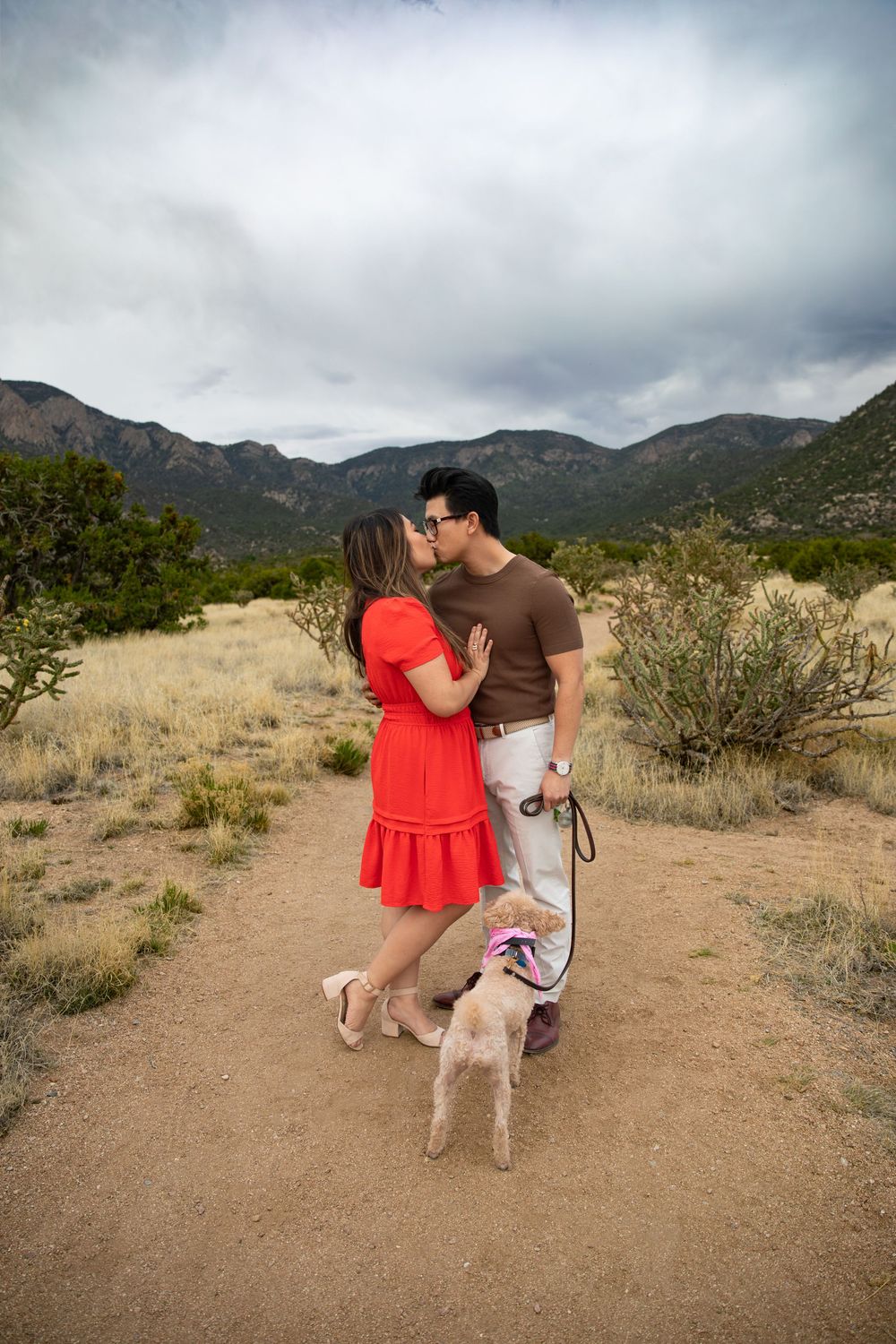 Couple kissing in desert landscape with mountains, woman in red dress, man in brown shirt, small dog on leash.