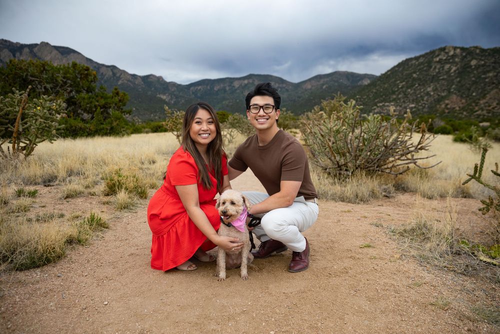 Couple in red dress and brown shirt kneeling with small dog in pink bow, desert mountain landscape backdrop.