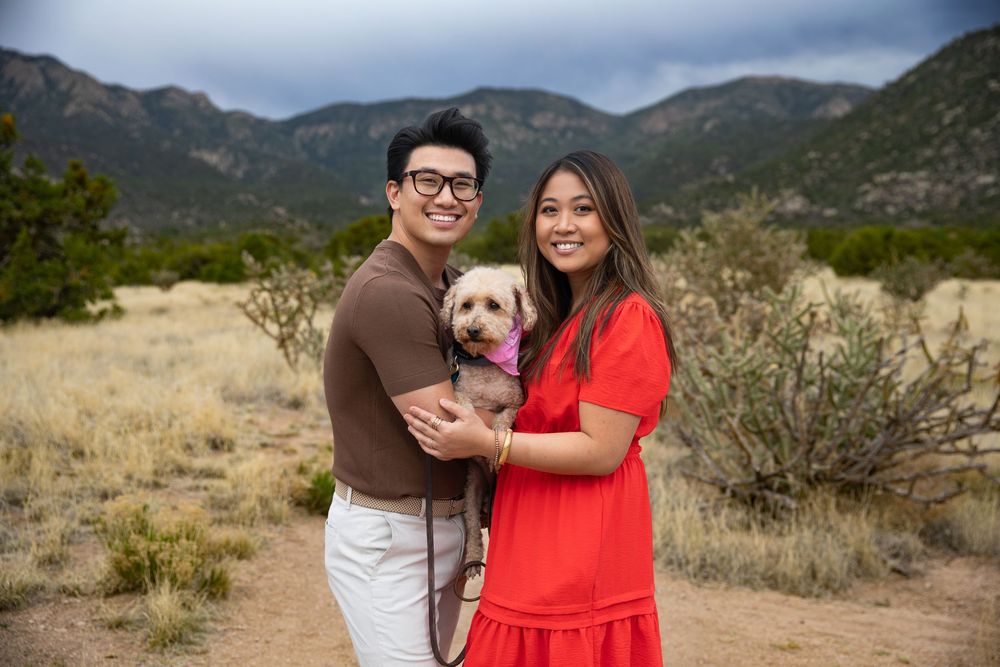 Couple smiling outdoors holding a small dog with mountain landscape background in a desert setting.