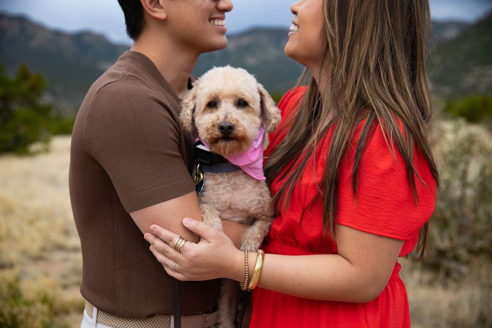 Couple in brown and red outfits holding a fluffy dog wearing a pink bandana outdoors with mountains in background.
