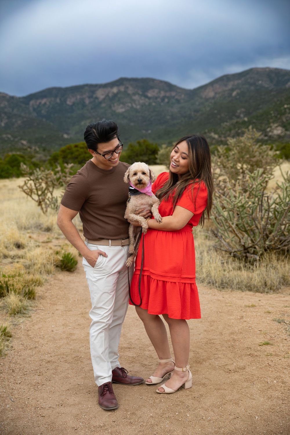 Couple in desert landscape holding small dog, man in brown shirt, woman in red dress, mountains in background.