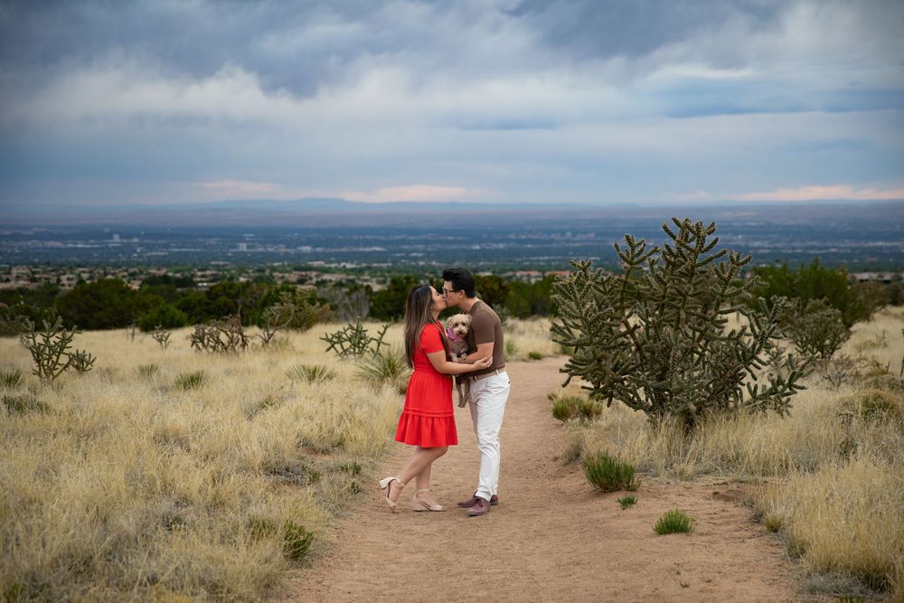 Couple kissing on a desert trail in red dress and white pants with vast valley and stormy sky backdrop.