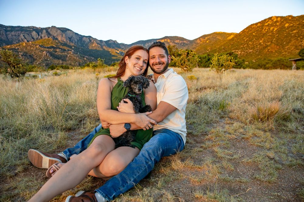 A couple sits together in tall grass at sunset with mountains and warm lighting in the background.