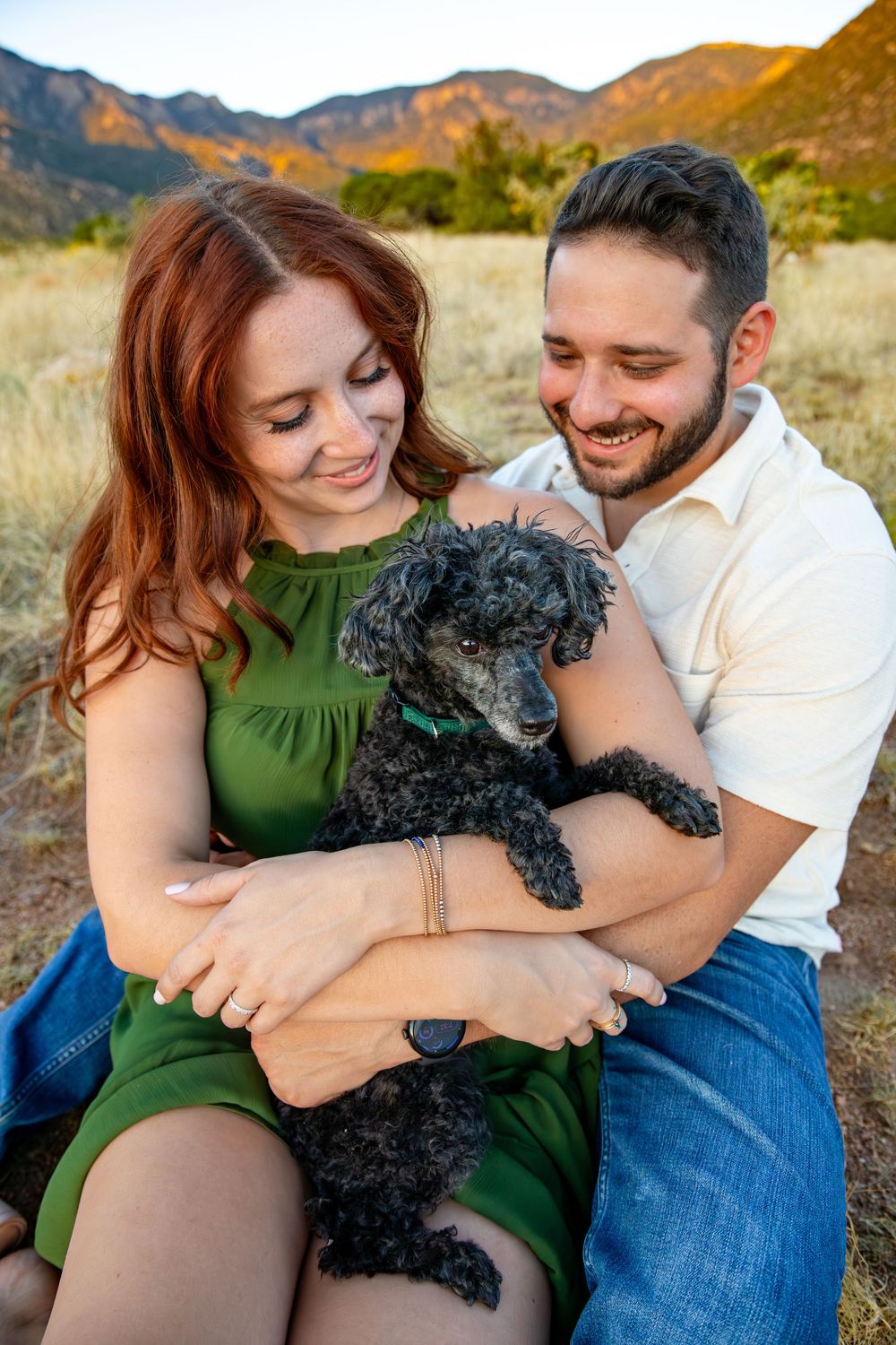 Couple cuddling with their black poodle dog while sitting outdoors in front of mountain scenery at sunset.