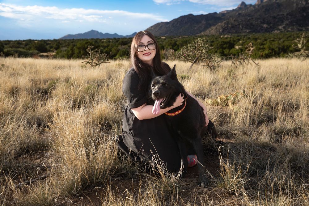 Woman in black outfit hugging a black dog in a dry grassy field with mountains in the background.