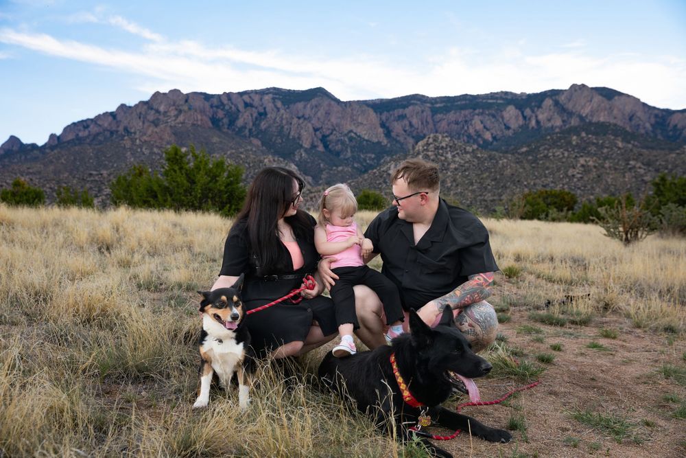 Family with toddler and husky dog sitting in desert field with Sandia Mountains in background.