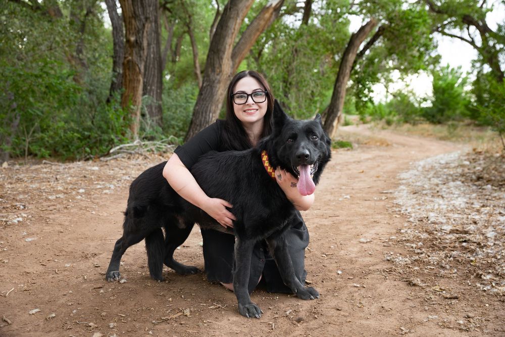 Woman with glasses poses with large black German Shepherd dog on a dirt trail surrounded by trees.