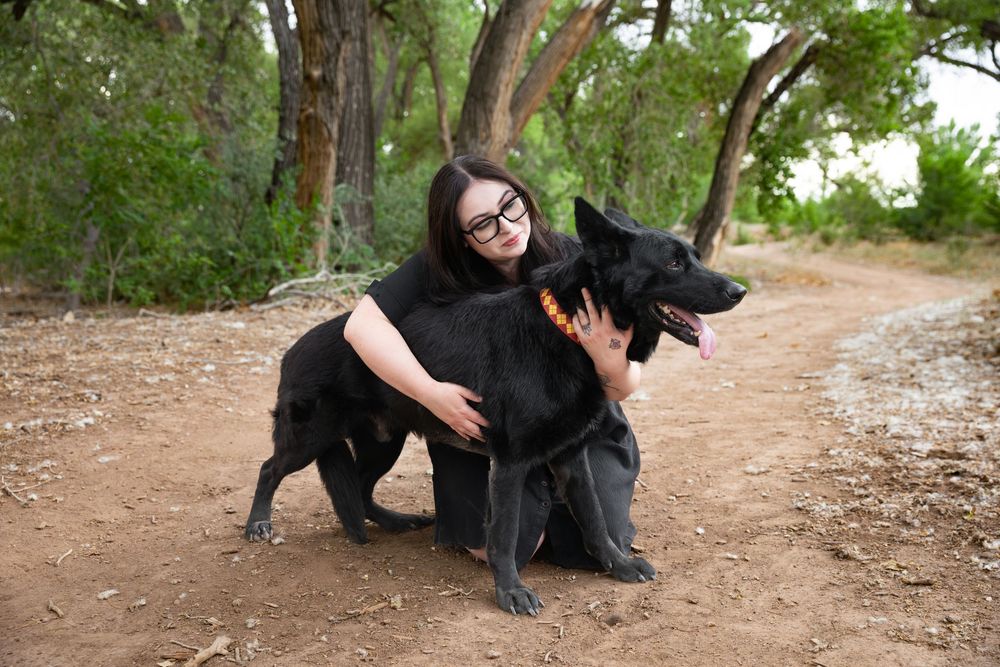 Woman with glasses hugging a large black German Shepherd dog on a dirt path surrounded by trees.