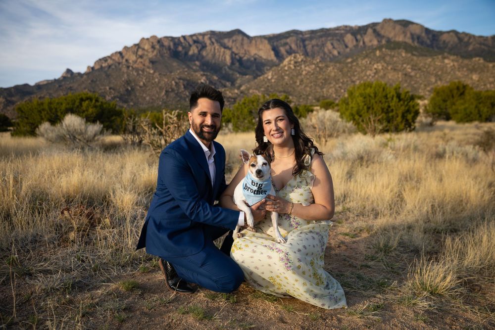 Couple in formal attire kneeling with their Jack Russell Terrier dog in a desert field with mountains behind.