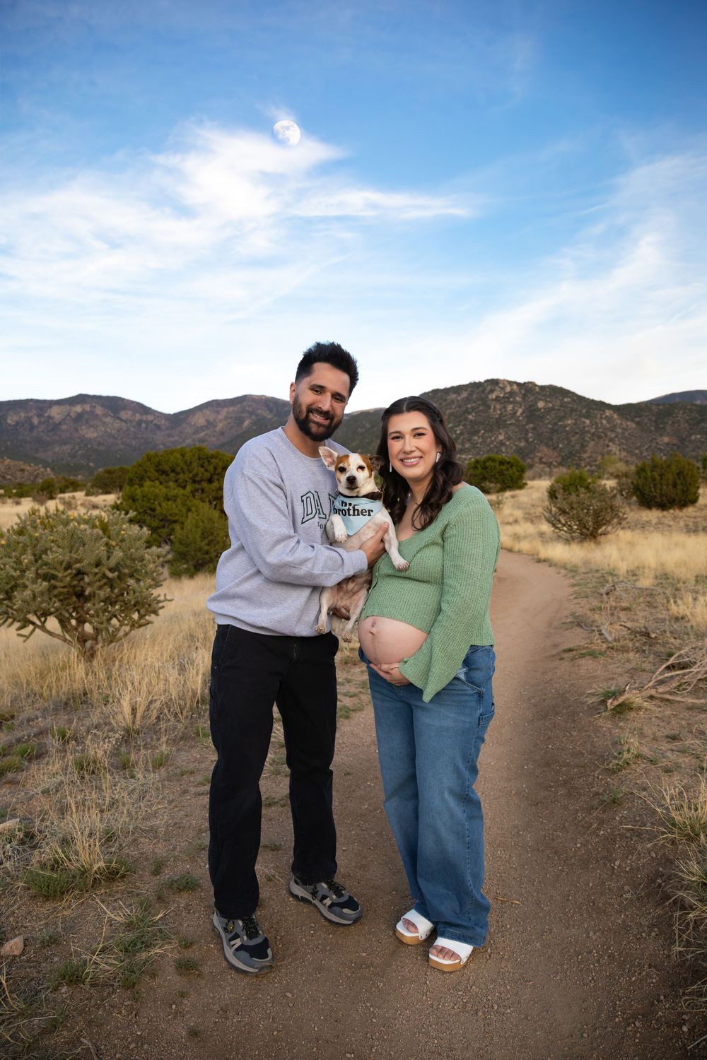 Pregnant couple holding small dog on desert trail with mountain views in New Mexico.