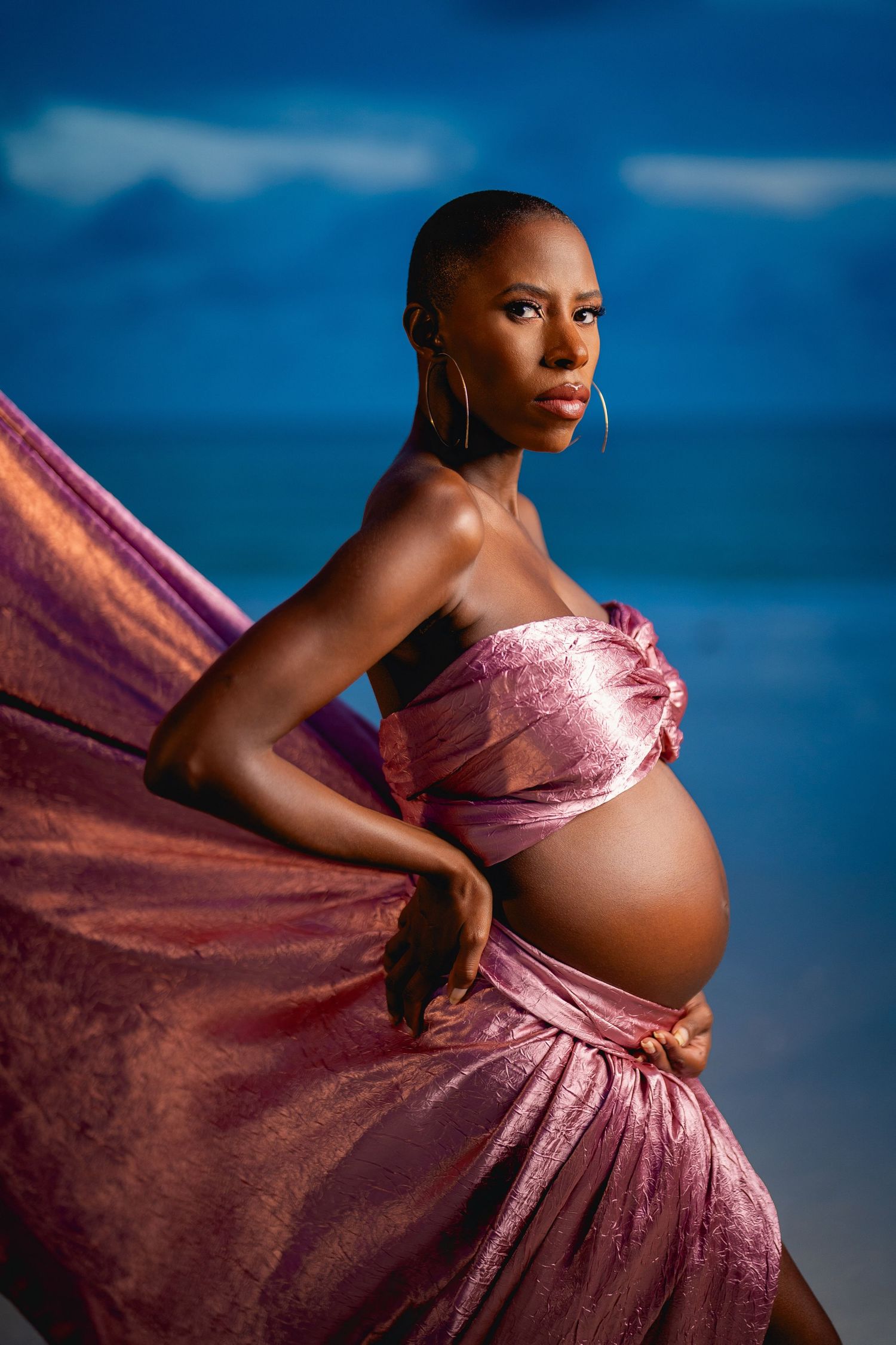 A pregnant person poses in a flowing pink silk maternity gown against a deep blue sky background.