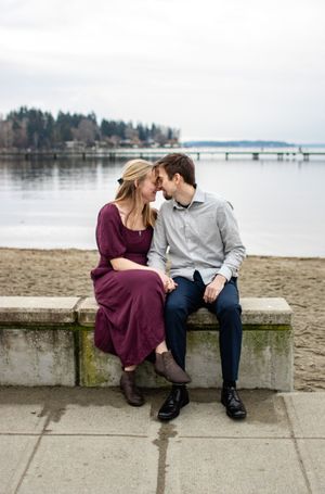 A couple sits close together on a stone bench by the waterfront, touching foreheads romantically on a cloudy day.