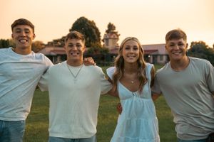 Four smiling teens stand together arms around each other at golden sunset outdoors.
