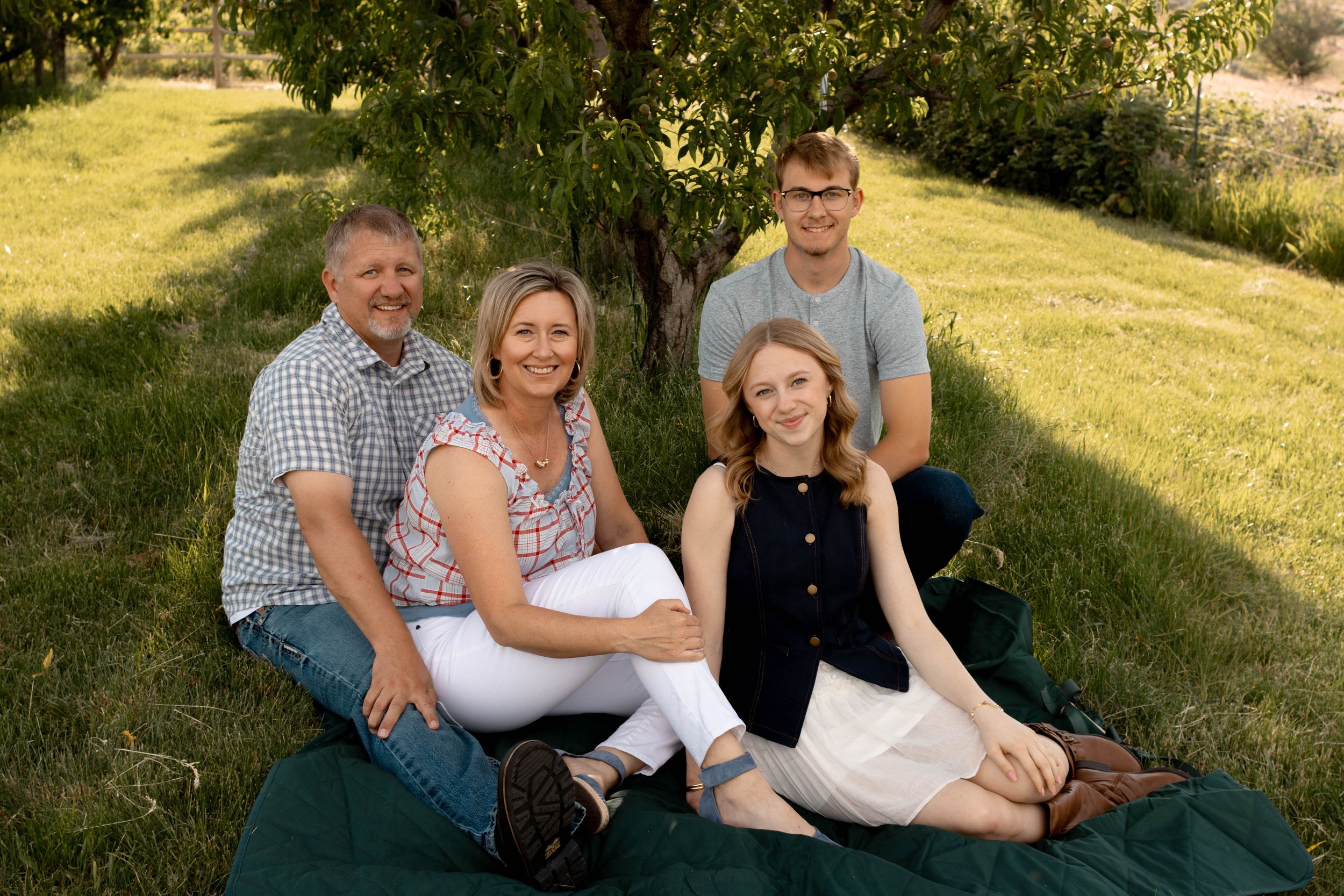 Family of four smiling together on a green blanket under a tree in a lush garden setting.
