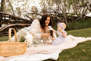 Mother and baby girl enjoying a picnic on a white blanket in a lush green park.