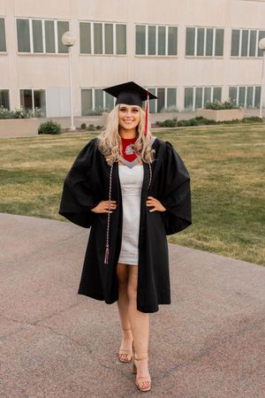 Smiling blonde woman in black graduation cap and gown standing outside a university building.