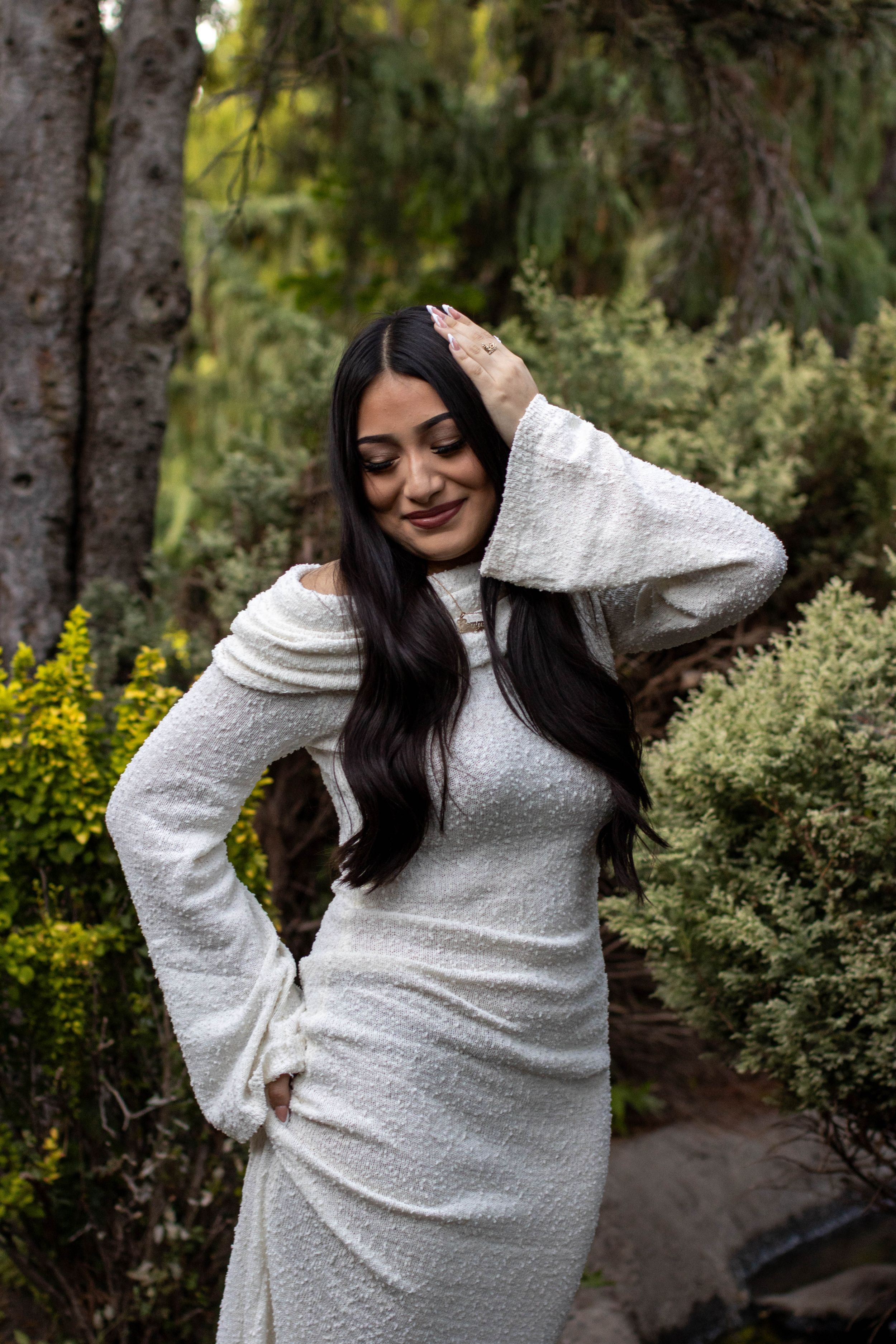 Woman in white off-shoulder dress poses gracefully outdoors among lush green trees and shrubs.