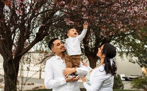 Happy family with parents holding toddler reaching up toward pink cherry blossom tree branches.