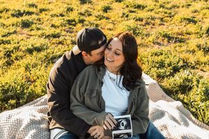 Couple sitting on a blanket outdoors holding an ultrasound photo as man kisses womans cheek.