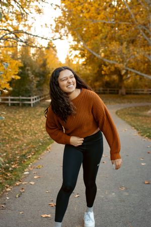 Young woman in rust sweater and black leggings laughing on a tree-lined path surrounded by golden autumn foliage.
