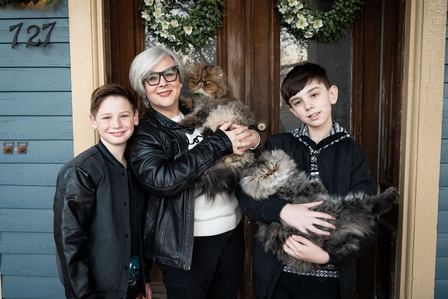 Three people and two fluffy cats pose together in front of a blue wooden door.