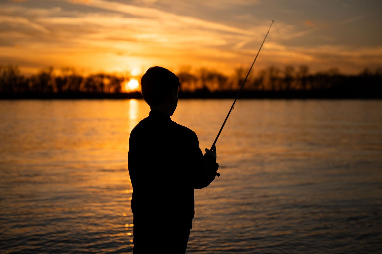A silhouette holds a fishing rod at sunset by a calm lake, with golden light reflecting on the water's surface.