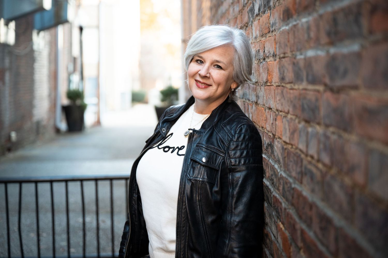 A person in a black leather jacket and white top stands next to a brick wall on a city street.