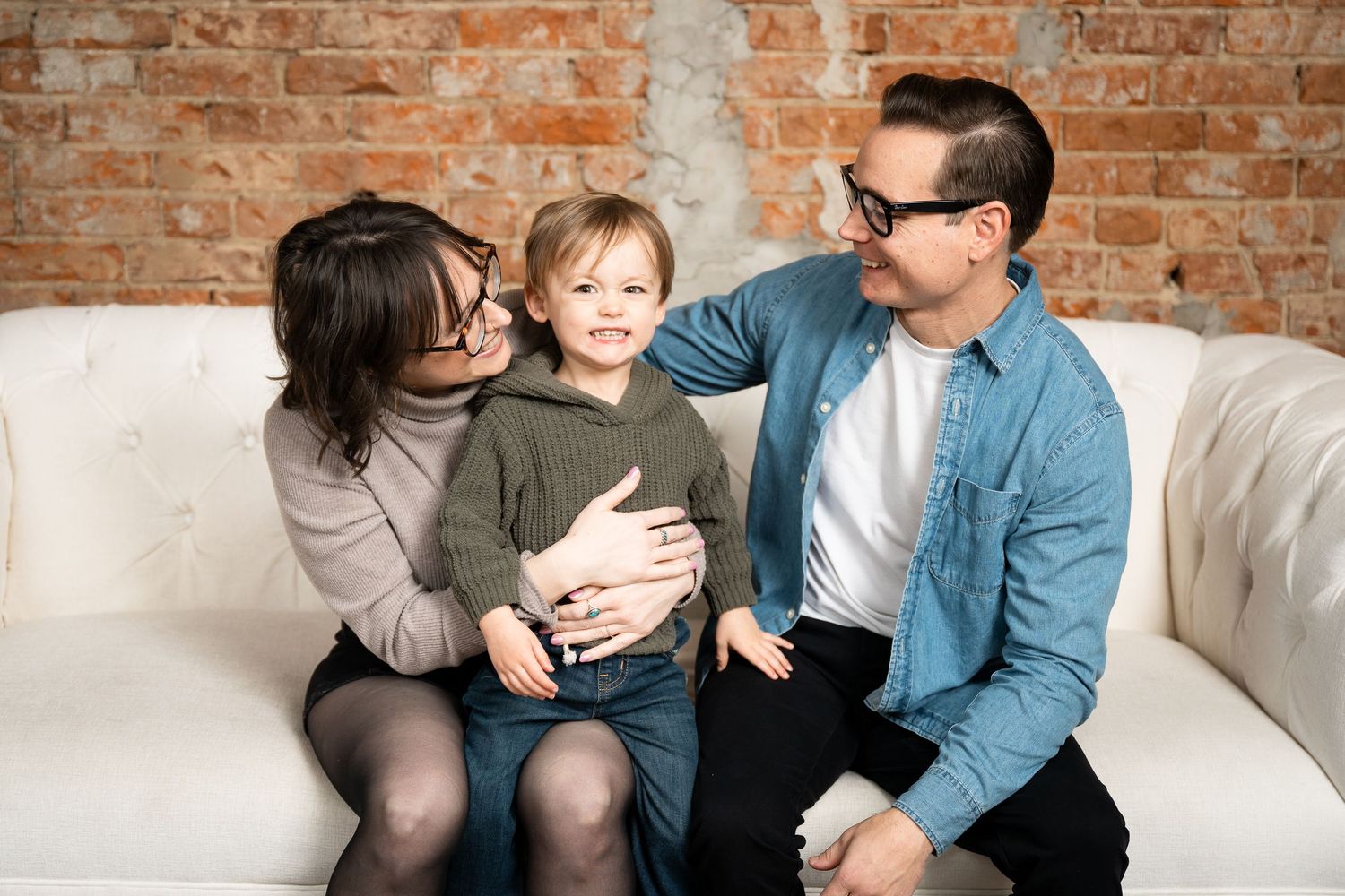 Three people sit together on a white couch against an exposed brick wall in a warm family moment.