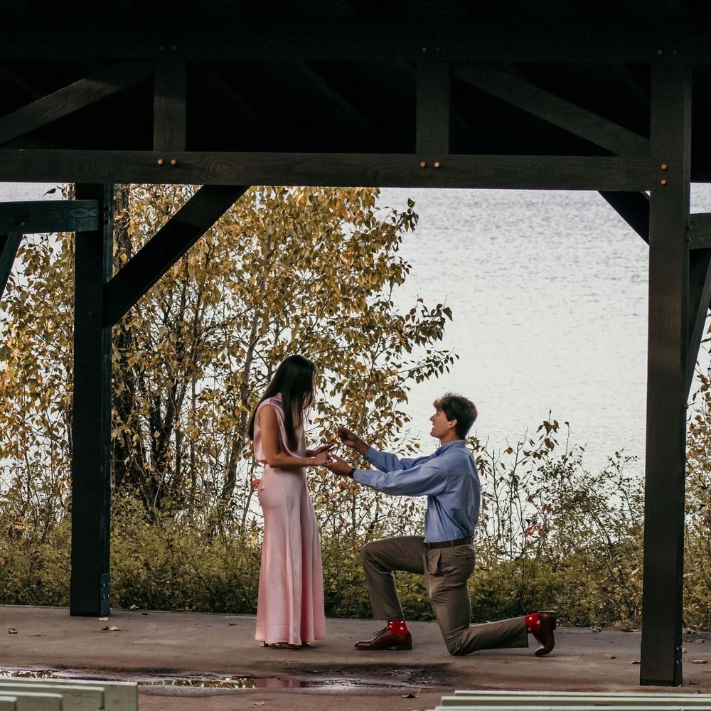 Man kneeling on one knee proposing to woman in pink dress under wooden pavilion near lake in autumn.