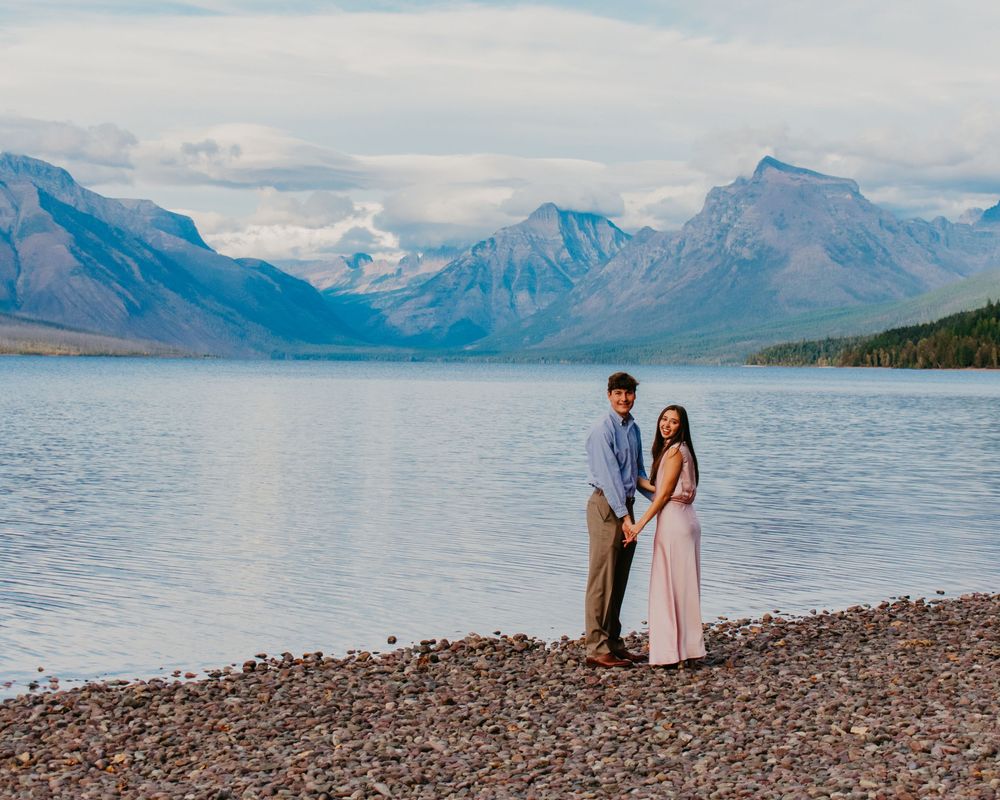 Couple holding hands on rocky lakeshore with stunning mountain backdrop at Glacier National Park.