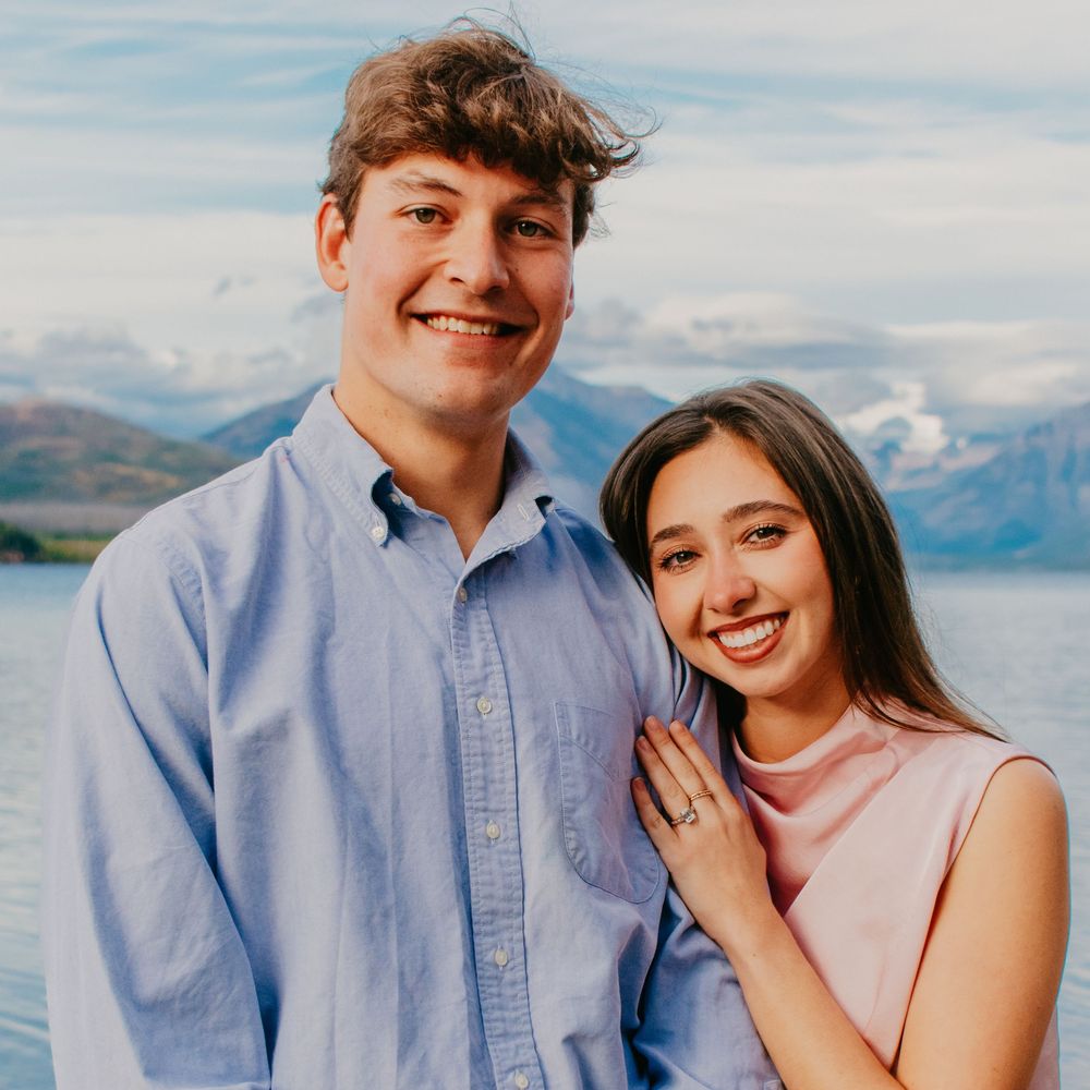 A smiling couple poses outdoors near a mountain lake, woman wearing an engagement ring, man in blue shirt.