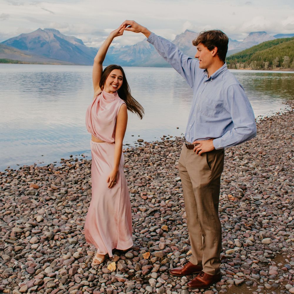 Couple dancing on rocky lakeshore with mountains in background, woman in pink dress, man in blue shirt.