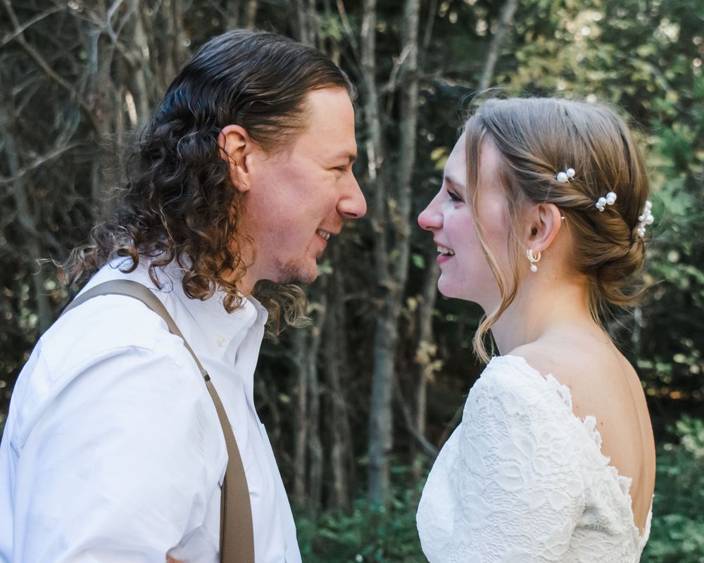 A bride and groom share a joyful moment face-to-face outdoors, she in lace gown, he in white shirt with suspenders.