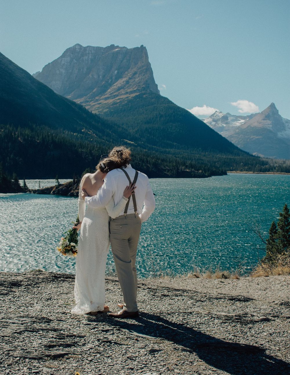 A bride and groom embrace while overlooking a turquoise mountain lake surrounded by peaks on their wedding day.