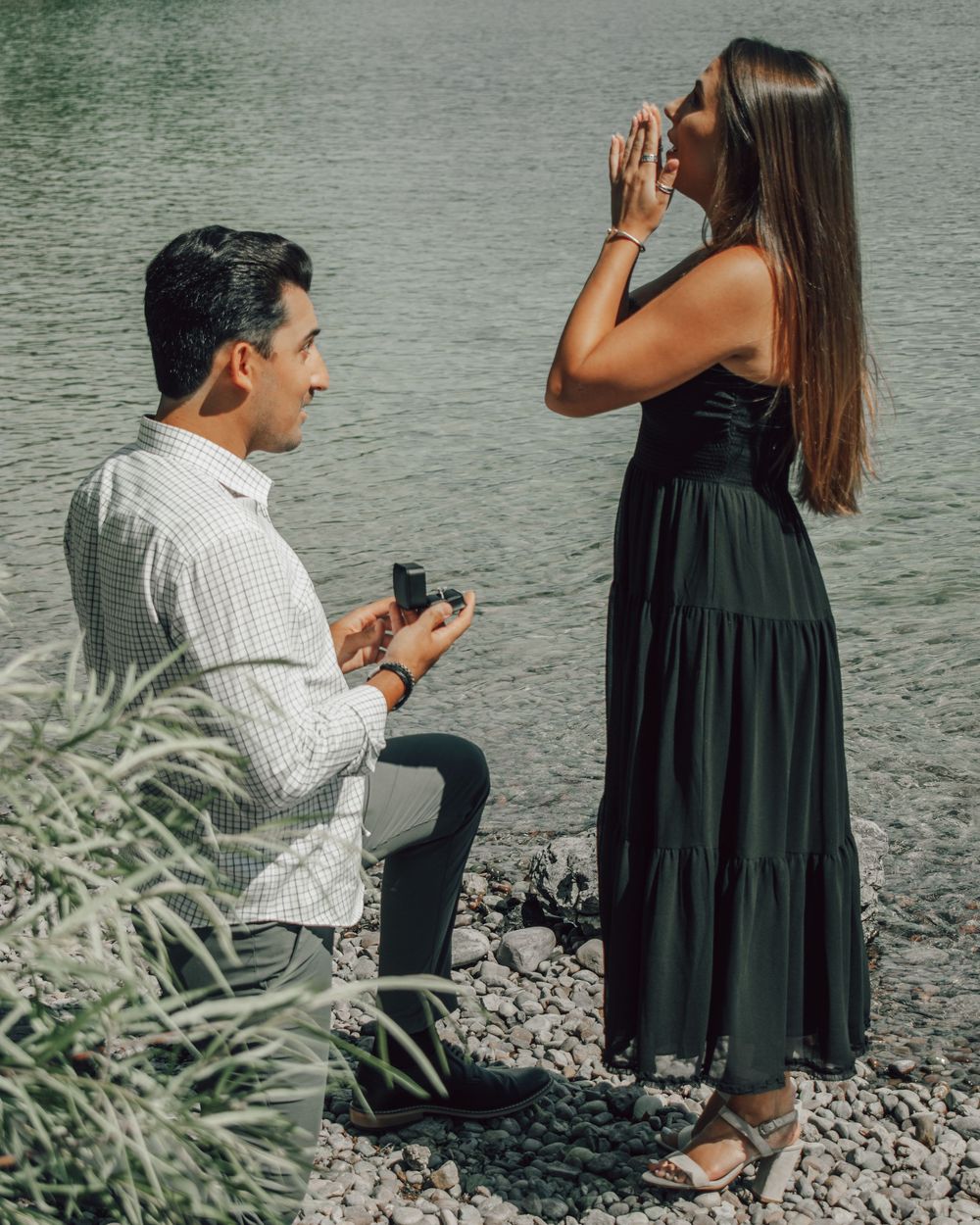 Man proposes to woman by a rocky lakeside, she reacts with joy, hands over mouth, wearing a black dress.
